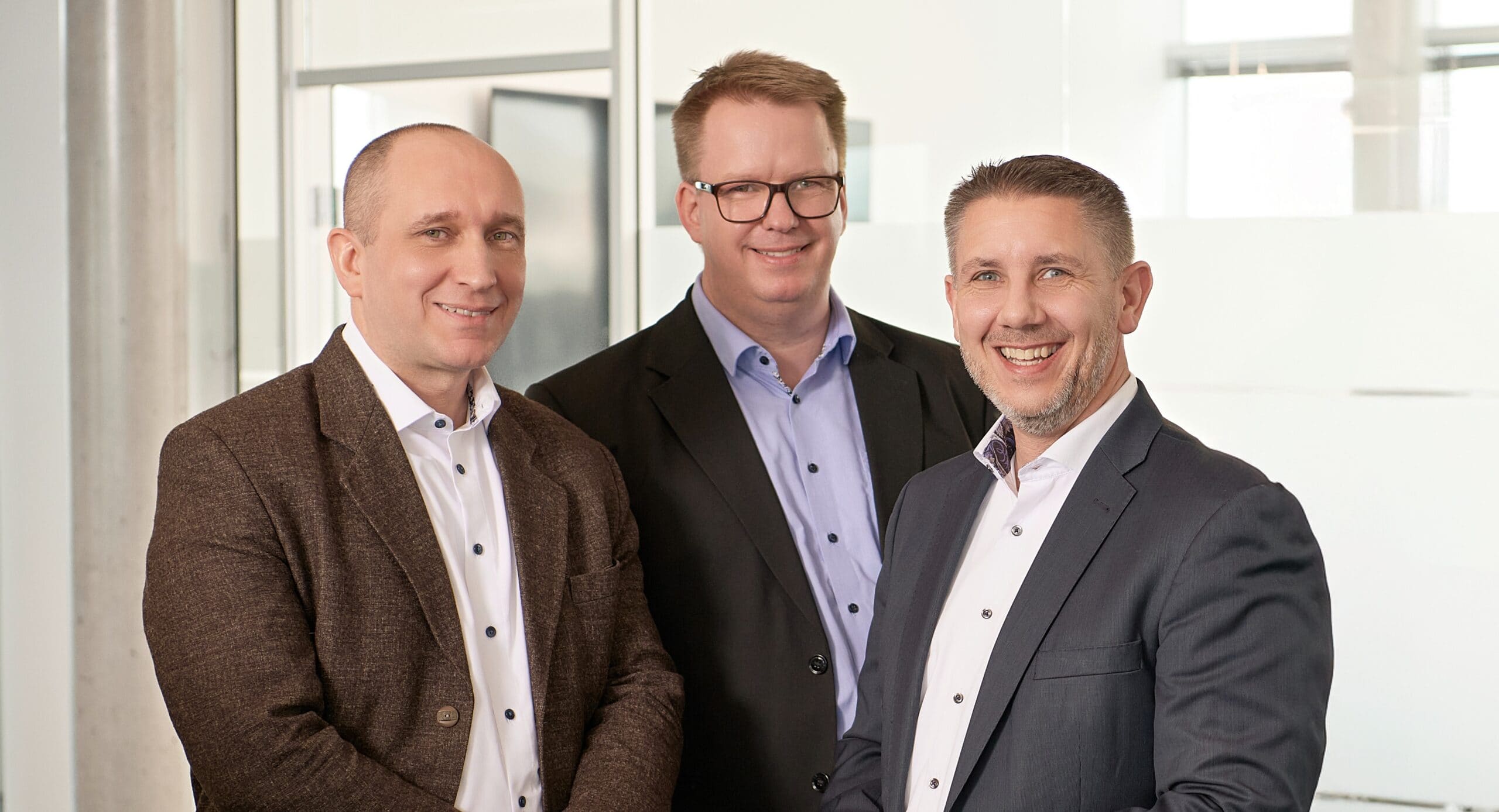 Drei Geschäftsleute in Anzügen vor einem modernen Bürogebäude.